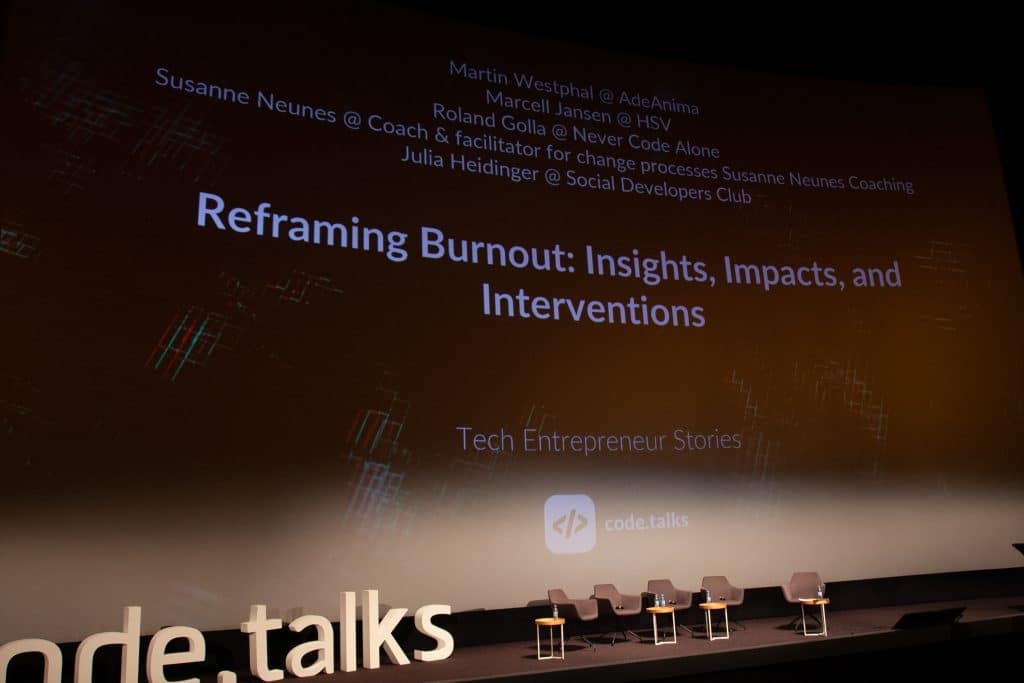 Code Talks 2023 Bühne mit Projektion des Programms auf eine Leinwand. Zu lesen sind die Namen der Teilnehmer sowie der Titel des Panels: "Reframing Burnout: Insights, Impacts and Interventions"
