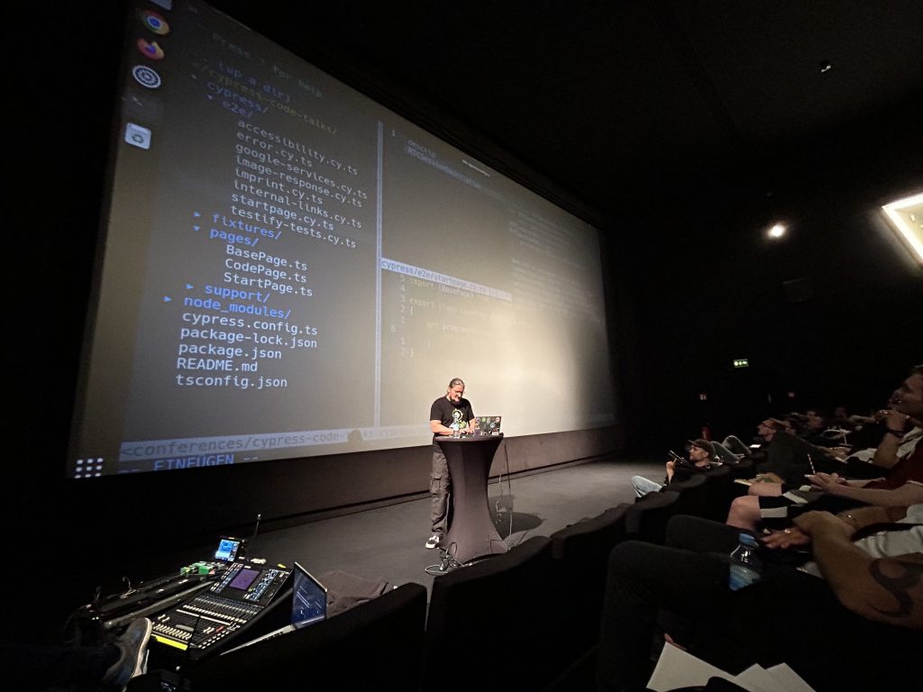 Roland Golla stehend an einem Stehtisch mit Laptop, auf der Bühne der code.talks. Hinter ihm wird Code auf eine Leinwand projeziert.